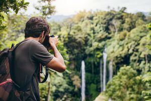 Hombre con mochila de pie frente a la cascada y tomando una foto. Excursionista macho fotografiando una hermosa caída de agua en el bosque