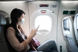 Young woman sitting using phone on the aircraft seat wearing face mask