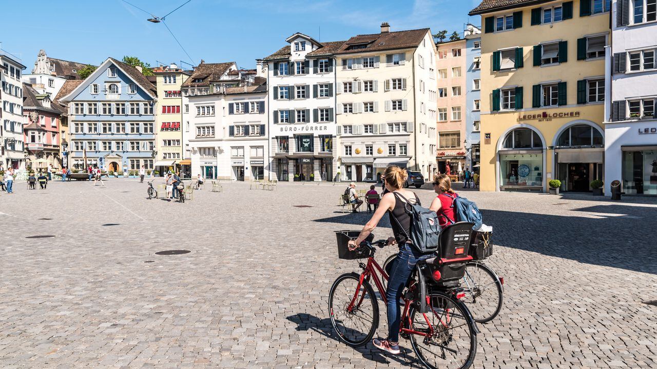 Dos señoritas andan en bicicleta por una plaza de la ciudad de Zúrich, la capital de Suiza. Otras personas caminan por los senderos pasando por las tiendas.