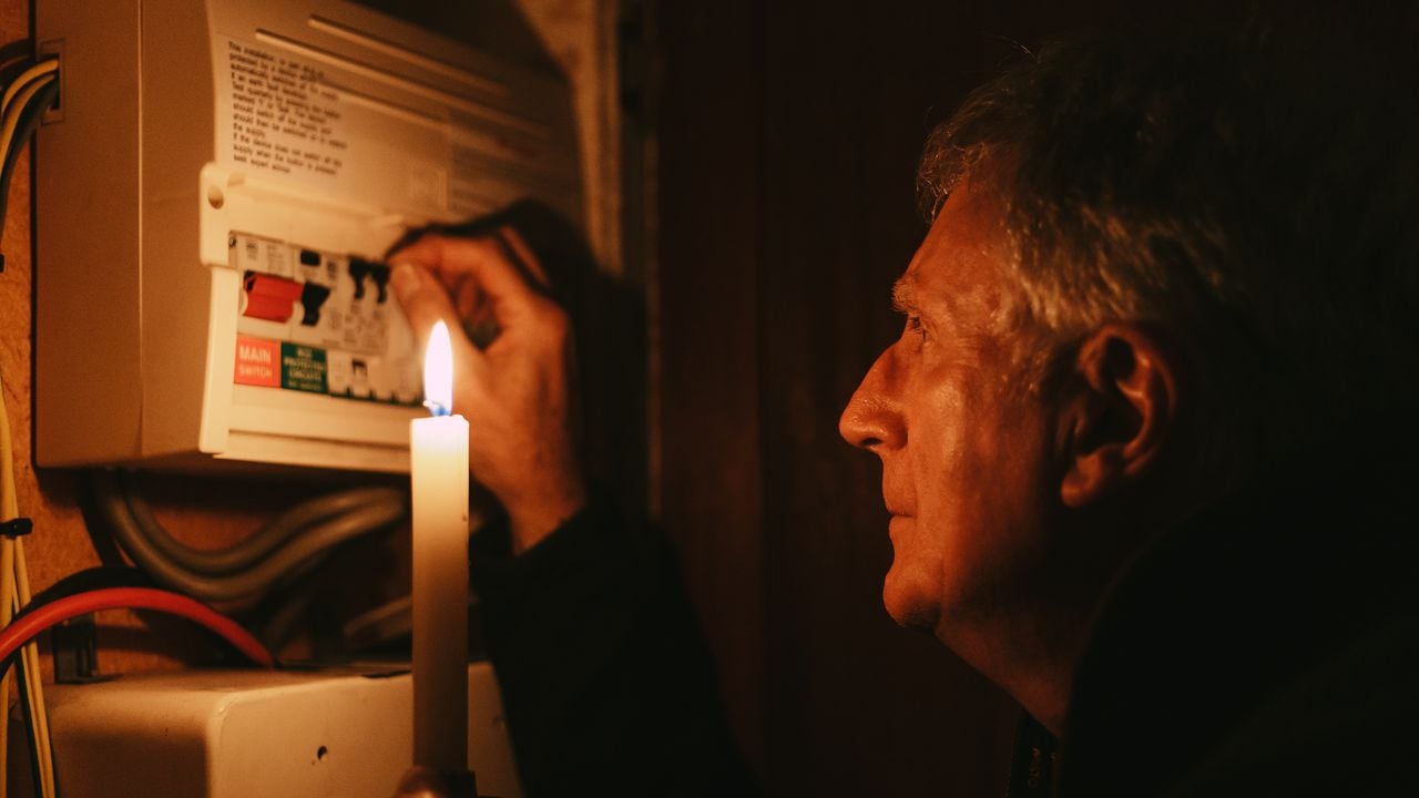 A senior man investigates his fuse box at home - by the light of a candle only - in a blackout during the energy crisis.