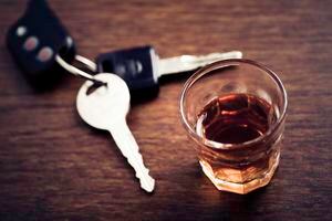 A shot glass full of dark colored alcohol on top of a bar table along with a set of car keys. Drinking and driving series. Also driving under the influence of alcohol.