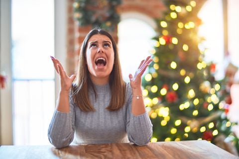 Young beautiful woman sitting at the table at home around christmas tree and decoration crazy and mad shouting and yelling with aggressive expression and arms raised. Frustration concept.