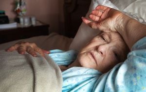 Anciana débil y febril en la cama cubriendo su frente con su brazo, tratando de descansar. Frascos de pastillas en el fondo.