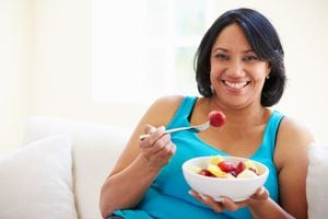 Mujer con sobrepeso sentada en un sofá comiendo un tazón de fruta fresca en casa mirando feliz