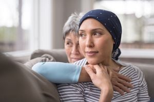 Una mujer étnica que lleva un pañuelo en la cabeza y lucha contra el cáncer se sienta en el sofá con su madre. Ella está en primer plano y su mamá está detrás de ella, con su brazo envuelto en un abrazo, y ambos están mirando por la ventana en un tranquilo momento de contemplación.