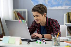 Varón adolescente sonriente charlando con amigos en las redes sociales sentado frente portátil