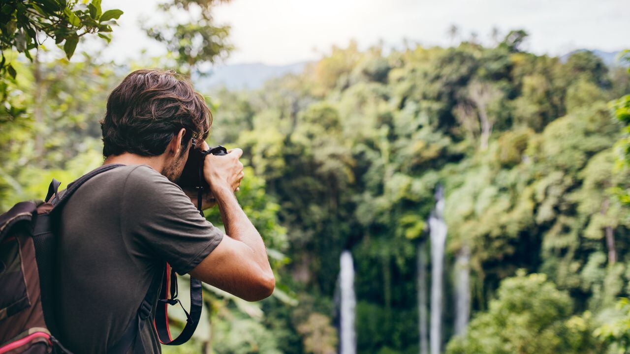 Hombre con mochila de pie frente a la cascada y tomando una foto. Excursionista macho fotografiando una hermosa caída de agua en el bosque