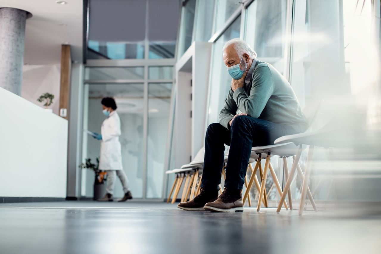Hombre mayor angustiado con mascarilla mientras está sentado solo en el pasillo del hospital. Copie el espacio.