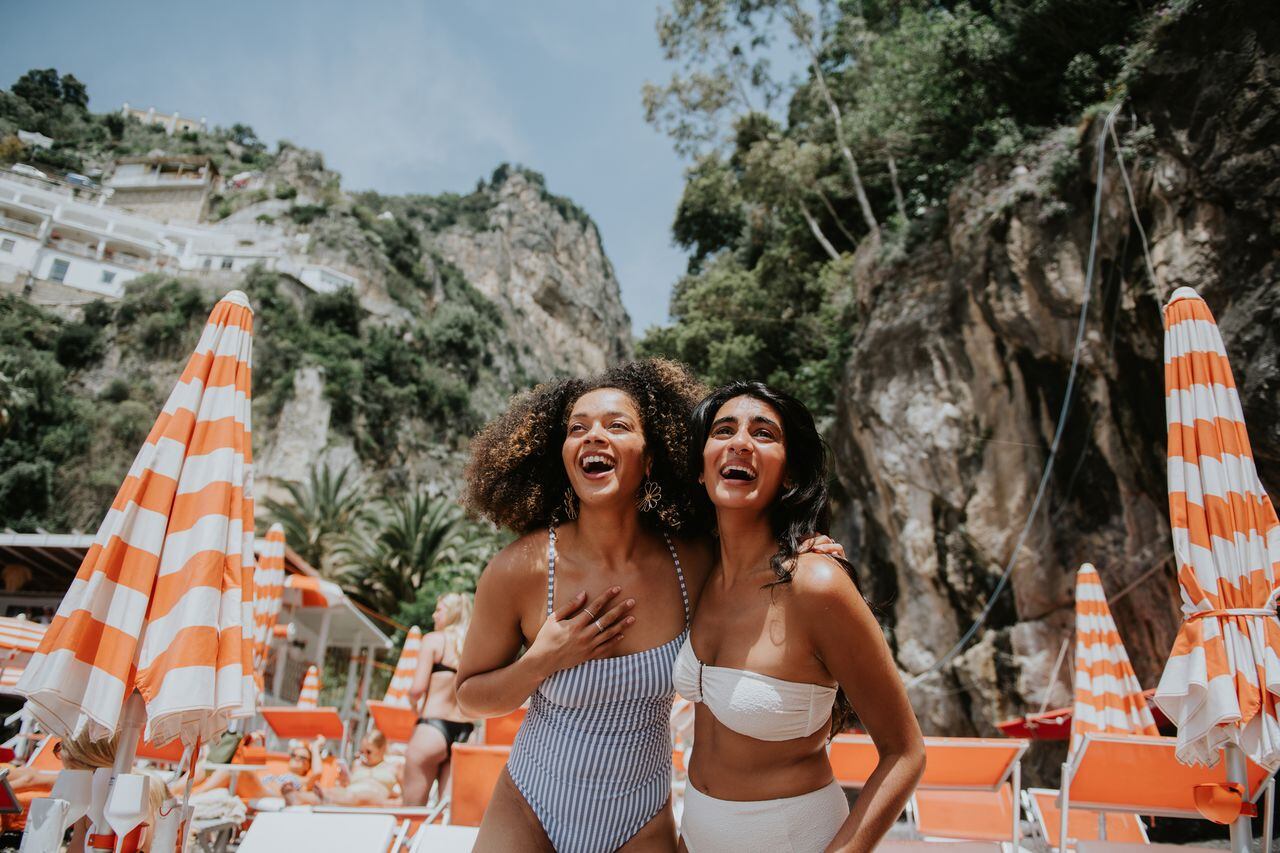Two woman look delighted to be holidaying together in Italy. Views of Positano are visible behind them. They are surrounded by striped beach umbrellas and sun loungers. They wear stylish swimwear.