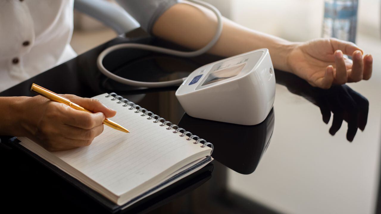 Woman taking blood pressure by using digital sphygmomanometer and record on empty white notebook or diary at home. Medical and healthcare concept.