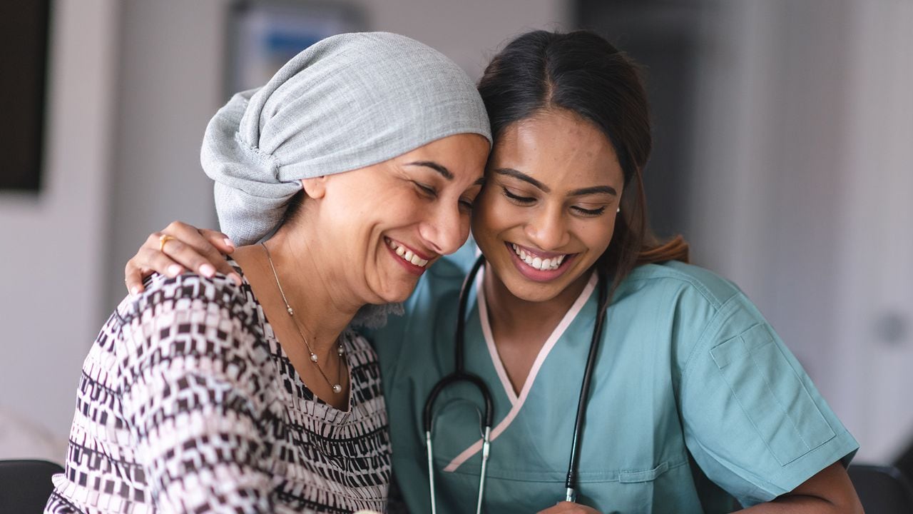 Retrato de una mujer india que lucha contra el cáncer con su médico. El doctor es una mujer mixta. Las dos mujeres están sentadas junto a la otra en el interior. Están abrazando y riendo.