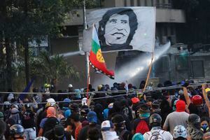 SANTIAGO, CHILE - JANUARY 03: Demonstrator carry a flag with face of popular singer Victor Jara whle a watter cannon fires against them during the first protest of the year against Chilean government on January 3, 2020 in Santiago, Chile. Chile's senate has announced today they will approve an increase of taxes on the richest individuals of the country as Chile's economy contracted 3.3% in November. Protests and social unrest arose on October 18 after a subway fare increase which developed in a social movement demanding improvements in basic services, fair prices and benefits including pensions, public health and education. (Photo by Marcelo Hernandez/Getty Images)