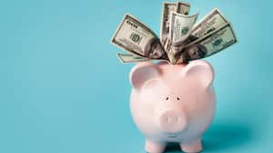 "Una alcancía rosa sonriente llena de billetes de 100 dólares, sobre fondo azul con espacio para copiar. También te puede interesar:"