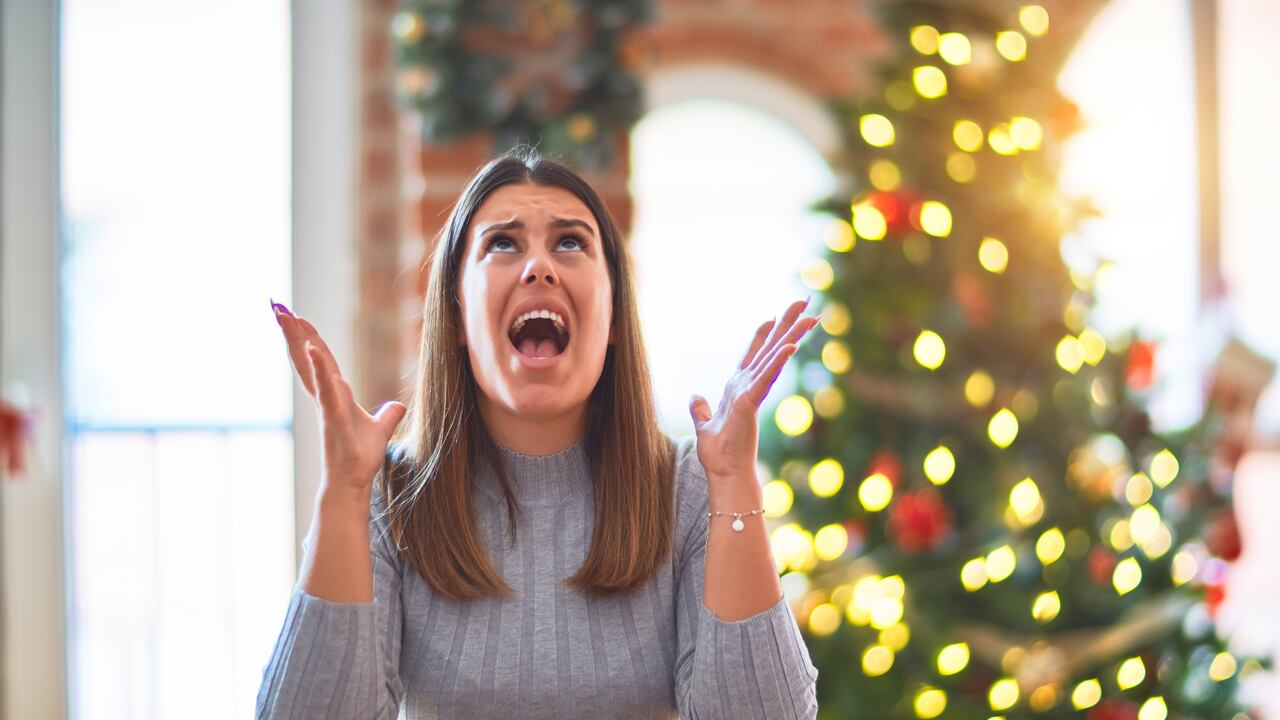 Young beautiful woman sitting at the table at home around christmas tree and decoration crazy and mad shouting and yelling with aggressive expression and arms raised. Frustration concept.