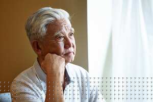 Retrato de un hombre mayor asiático triste sentado junto a la ventana con la mano en la barbilla.