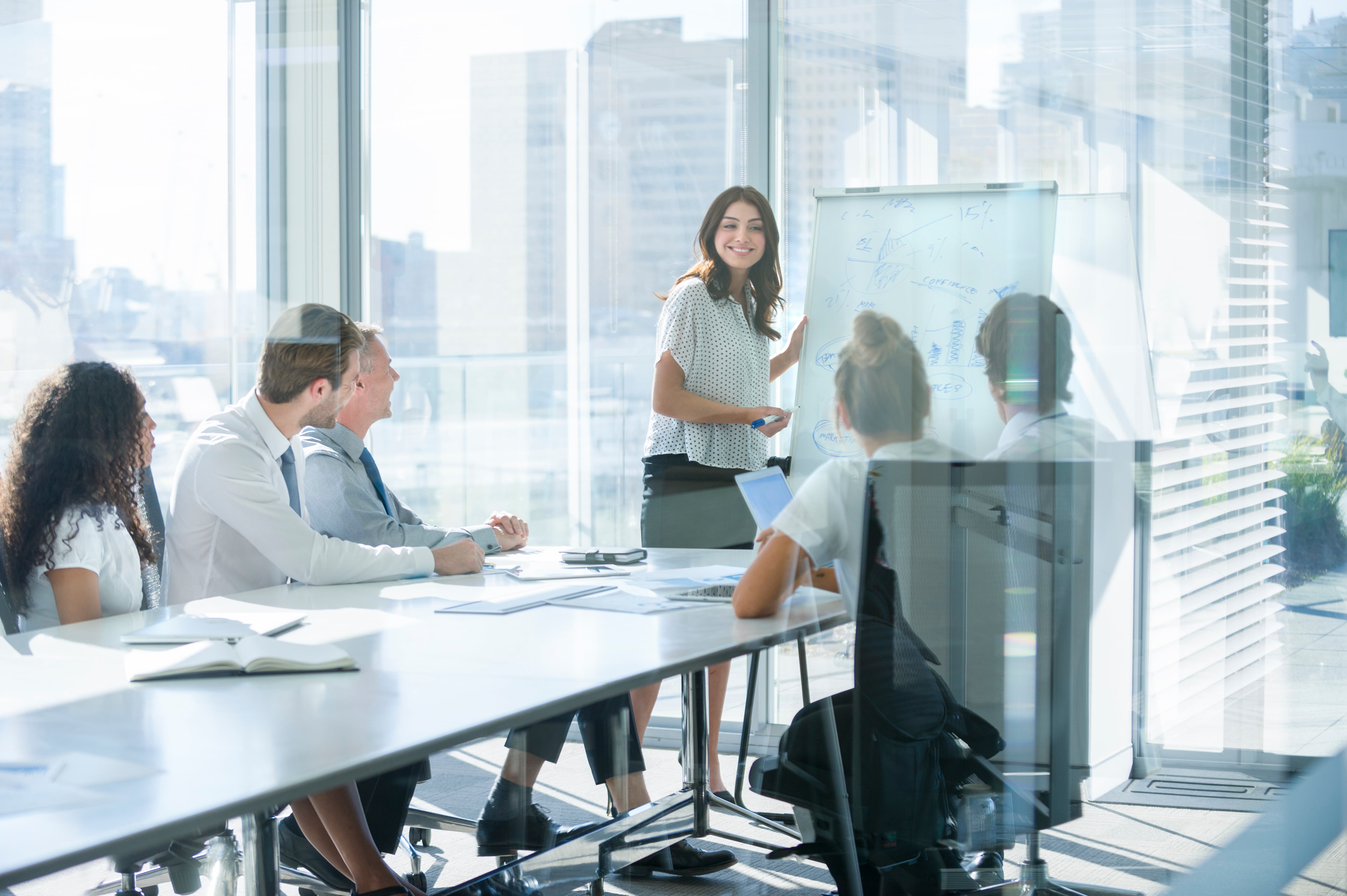 Mujer dando una presentación a su equipo. Ella está usando una pizarra con tablas y gráficos. Ella está hablando. Están en una sala de juntas de la oficina en la mesa con computadoras portátiles y papeles. Ella esta sonriendo. Hombres y mujeres del grupo.