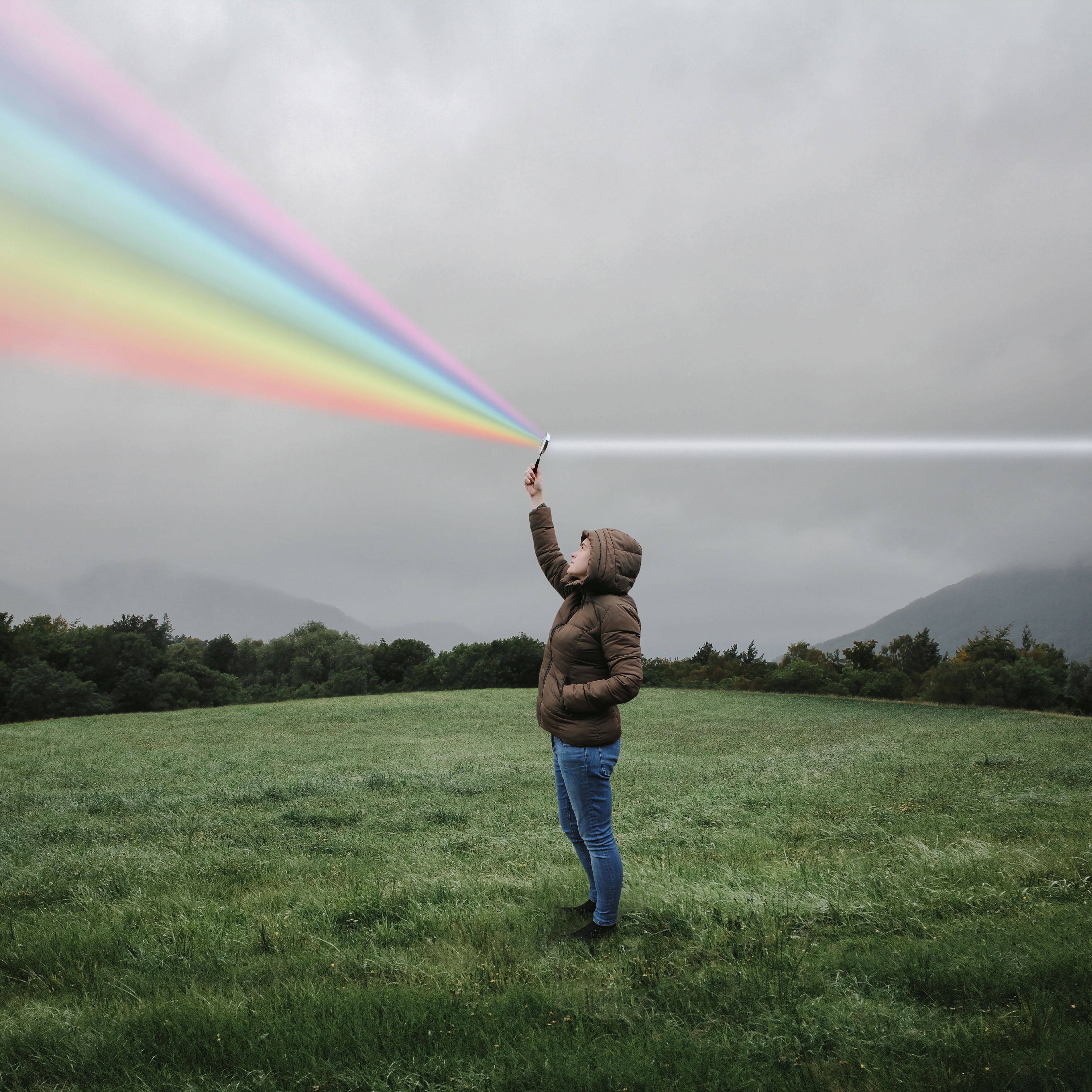 Woman standing on a cloudy day in a field holding up a magnifying glass shining a rainbow.