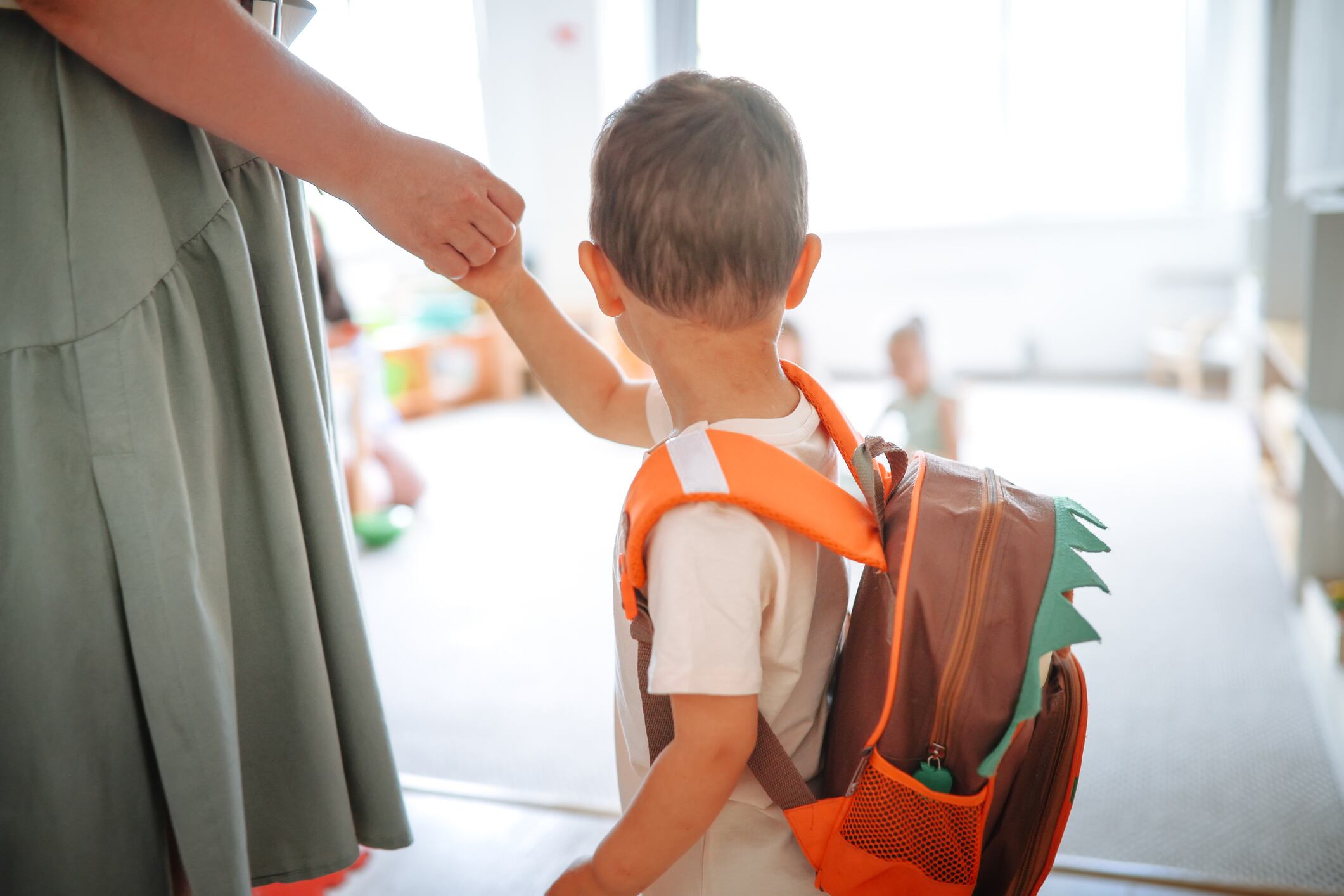 Asian toddler with a backpack holds mom's hand arriving at kindergarten, adapting to the children's group