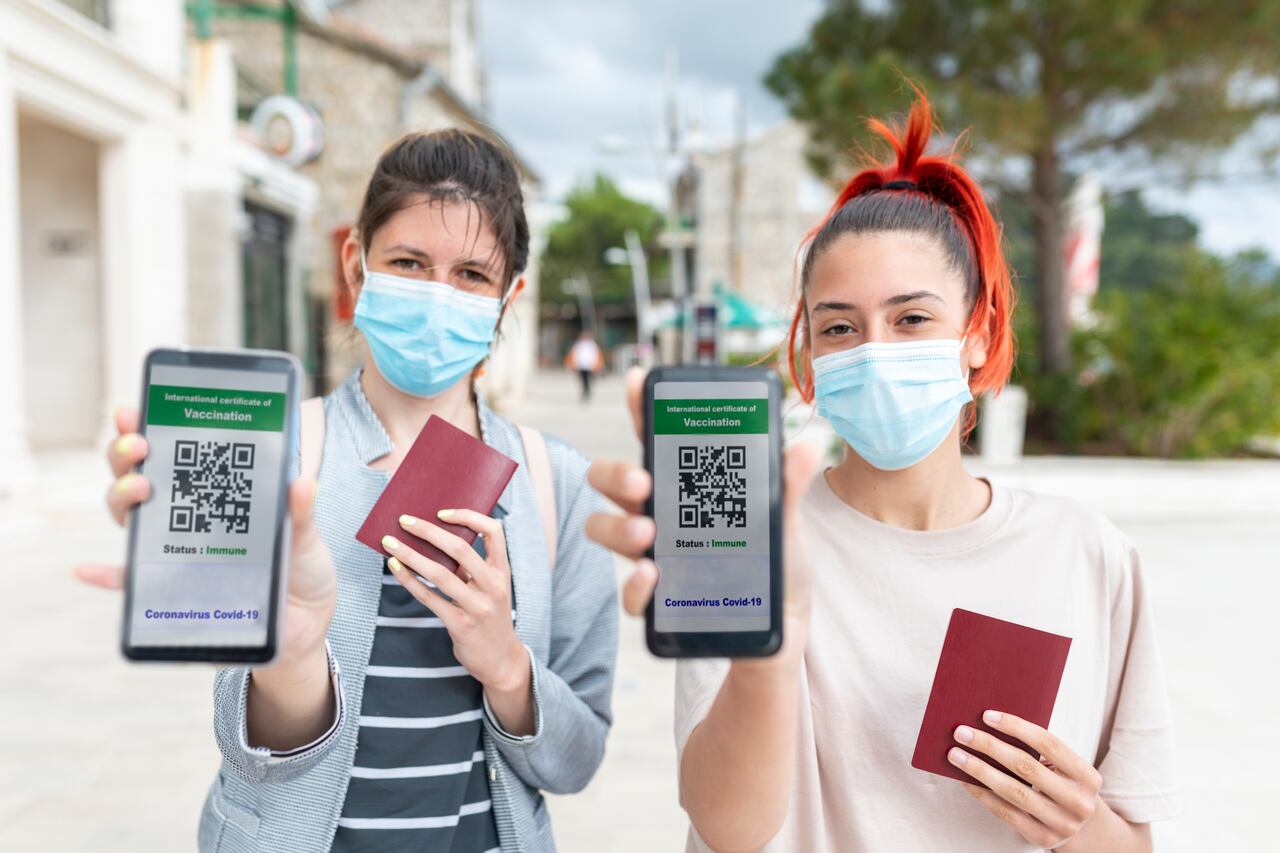 Dos mujeres jóvenes, turista caminando por una ciudad costera con máscaras en el rostro y pasaportes en la mano. Están mostrando una pantalla de teléfono inteligente con certificado electrónico de vacunación COVID19. Unas vacaciones tan esperadas