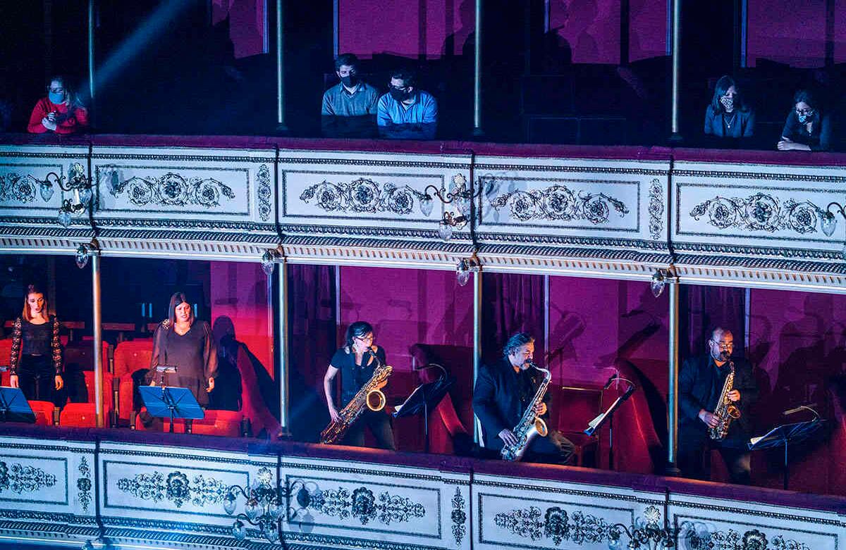 Algunas personas con mascarillas escuchan a los músicos de la Banda Sinfónica de Montevideo que actúan debajo de ellos en el teatro Solís en Montevideo, Uruguay, el jueves 13 de agosto. Cinco meses después de que todos los espectáculos de artes en vivo fueran cancelados debido a la pandemia, el teatro Solís reabrió para una actuación pública. Foto: Matilde Campodonico / AP  