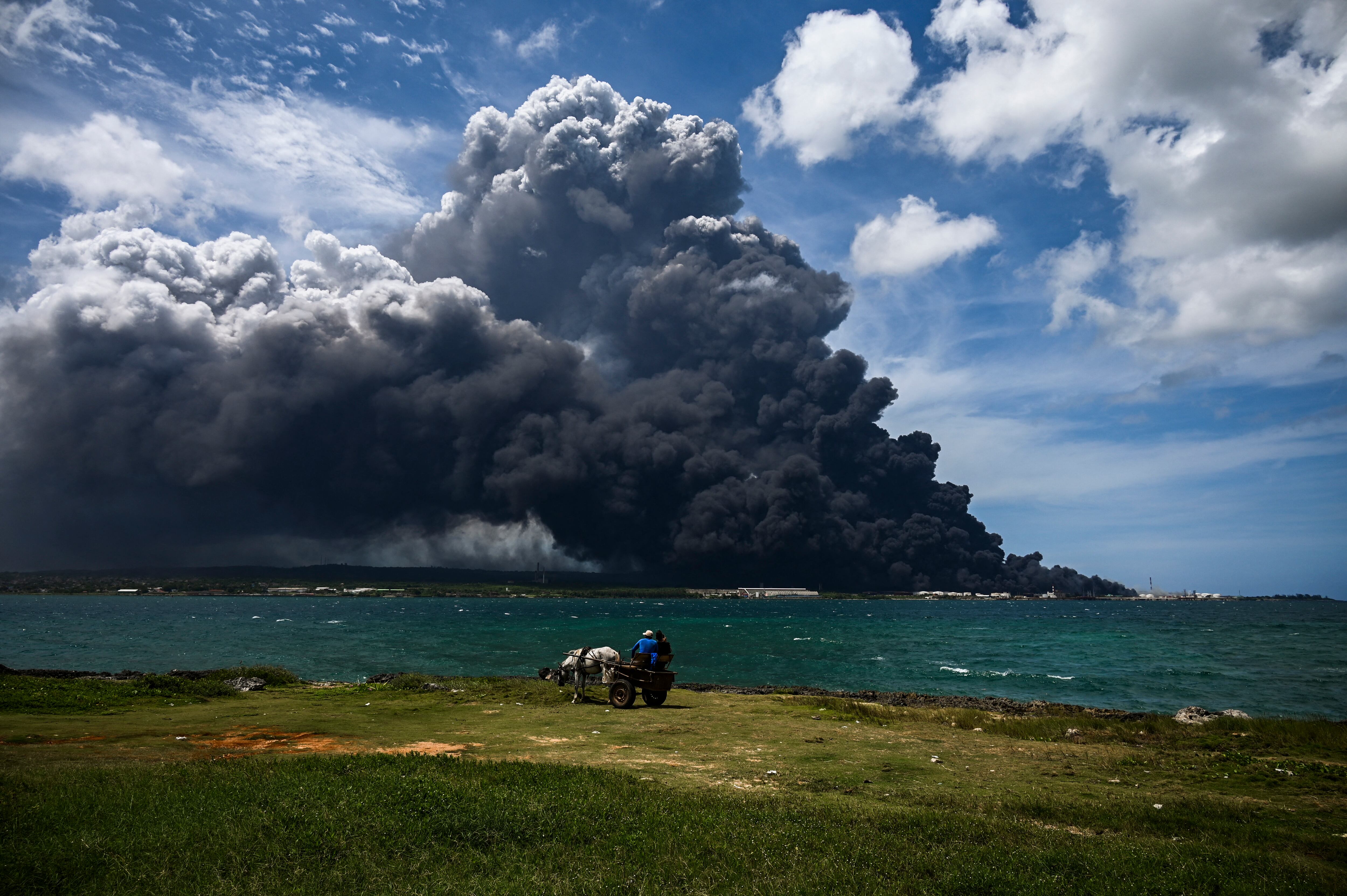 Las personas en un carro de caballos miran el humo negro del incendio masivo en un depósito de combustible en Matanzas, Cuba, el 8 de agosto de 2022. - Helicópteros se apresuraron a contener un incendio de varios días que derribó un tercer tanque en un depósito de combustible el lunes mientras continuaba la búsqueda de 16 bomberos desaparecidos. (Foto de YAMIL LAGE / AFP)