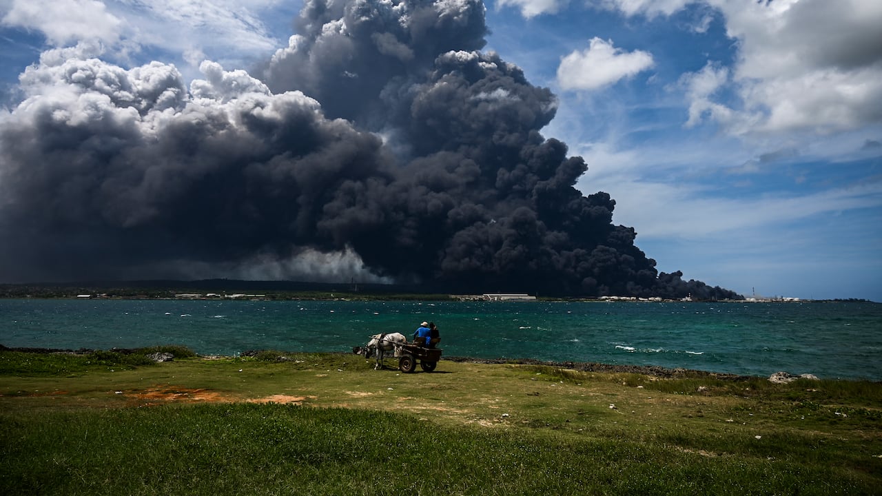 Las personas en un carro de caballos miran el humo negro del incendio masivo en un depósito de combustible en Matanzas, Cuba. Helicópteros se apresuraron a contener un incendio de varios días que derribó un tercer tanque en un depósito de combustible el lunes mientras continuaba la búsqueda de 16 bomberos desaparecidos.