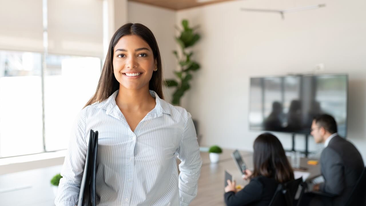 Portrait of hispanic businesswoman with a file standing in meeting room with colleagues disucssing in background