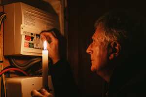 A senior man investigates his fuse box at home - by the light of a candle only - in a blackout during the energy crisis.