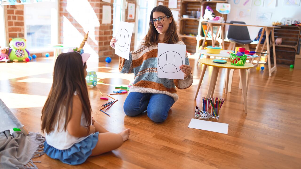 Hermosa psicóloga y niño pequeño haciendo terapia con emociones emoji alrededor de muchos juguetes en el jardín de infantes
