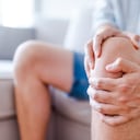 Man suffering from knee pain sitting sofa. A mature man massaging his painful knee. Man suffering from knee pain at home, closeup. Pain knee
