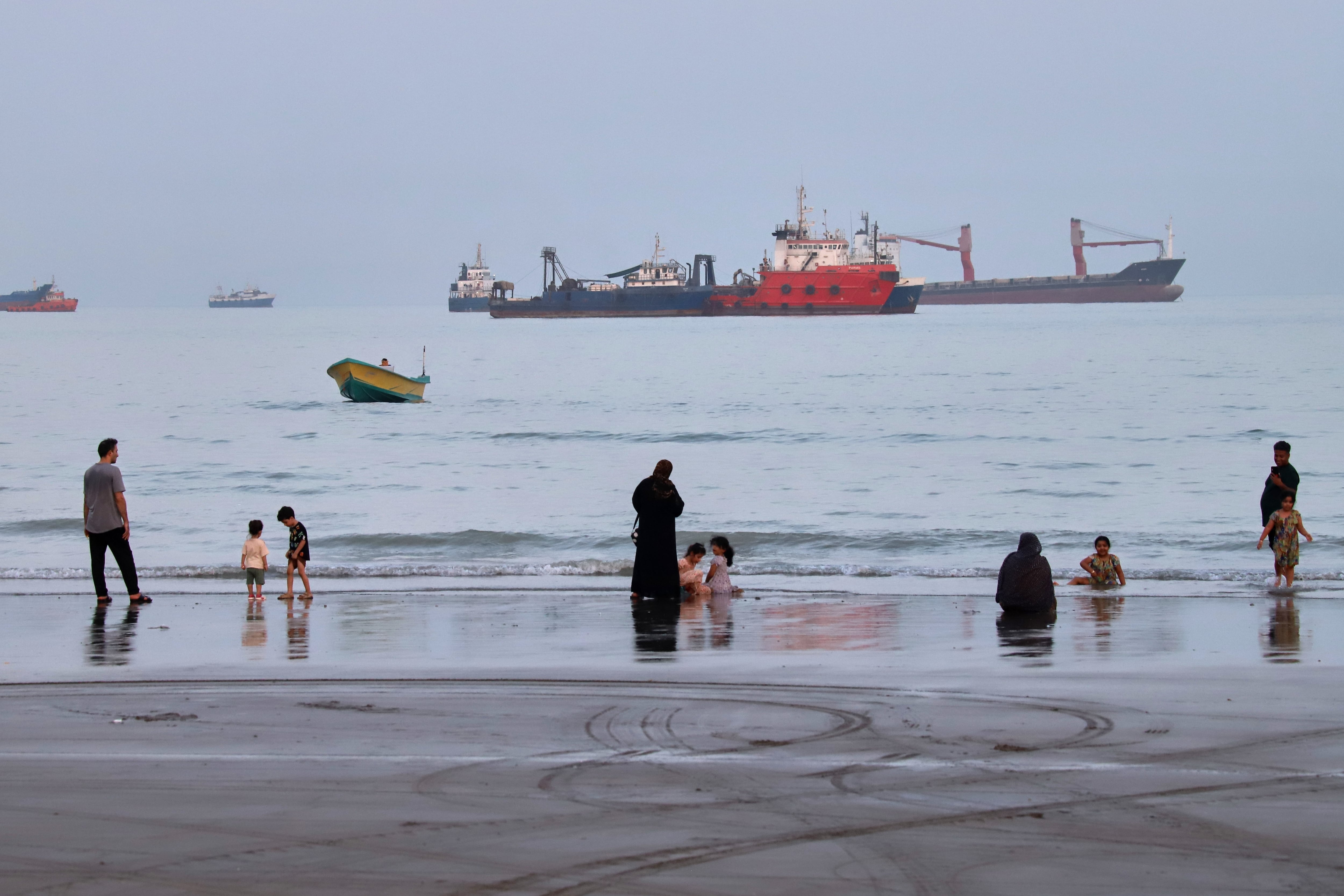 Gente pasa tiempo en la playa mientras se ven mercantes fondeados detrás en el estrecho de Ormuz, cerca de Bandar Abbas, Irán.