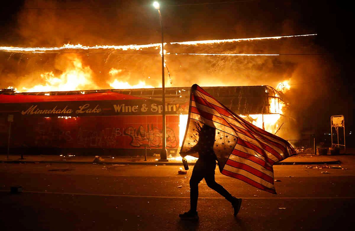 Un manifestante lleva la bandera de Estados Unidos al revés, una señal de angustia, junto a un edificio en llamas el jueves 28 de mayo de 2020 en Minneapolis. Las protestas por la muerte de George Floyd, un hombre negro que murió bajo custodia policial el lunes, estalló en Minneapolis por tercera noche consecutiva. (Foto AP / Julio Cortez)