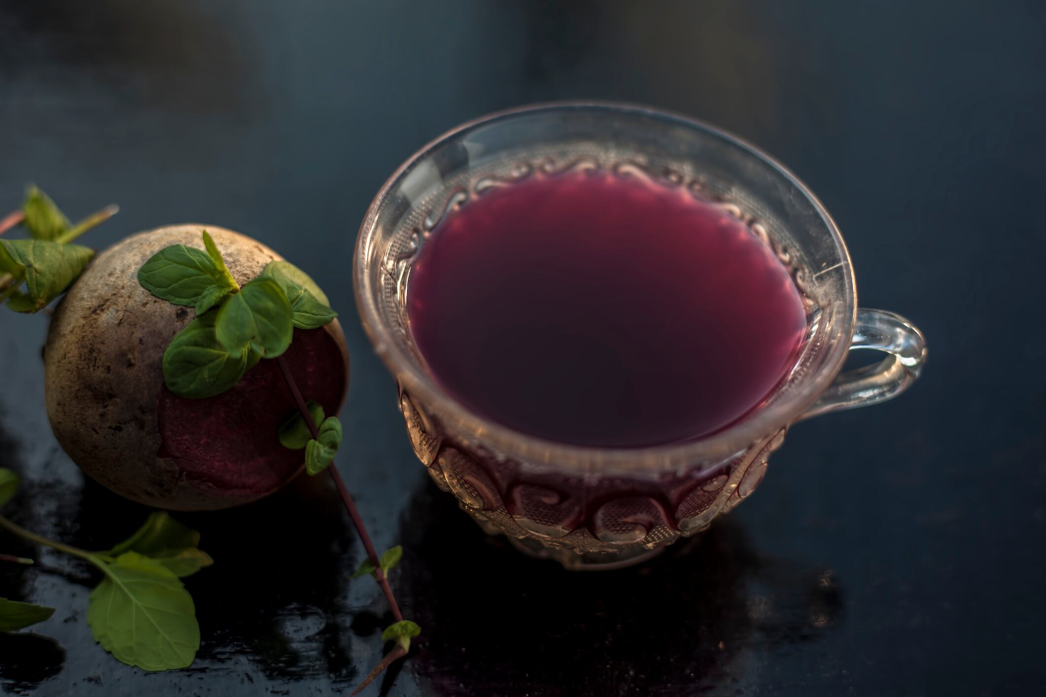 Best detoxify drink on a black glossy surface in a glass cup. Beetroot tea in a transparent glass cup on a black surface with a raw beet and some mint leaves. Horizontal shot with blurred background.