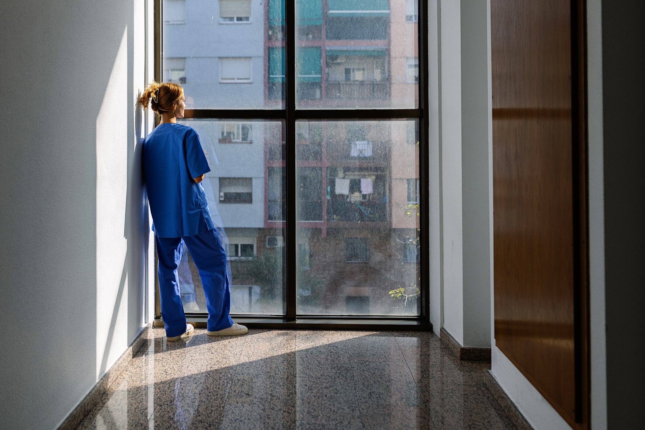 Vista trasera de una joven trabajadora de la salud vestida de azul mirando por la ventana del hospital por la mañana