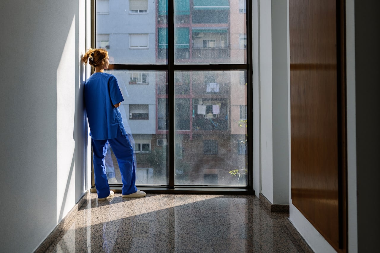Vista trasera de una joven trabajadora de la salud vestida de azul mirando por la ventana del hospital por la mañana