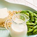 A variety of soy milk and soybean products. They are sitting in a soft aqua colored tray. There is a brick of firm tofu and a small pitcher of soy milk. On the right are some green soy beans and in back there is a small dish of shelled soybeans.