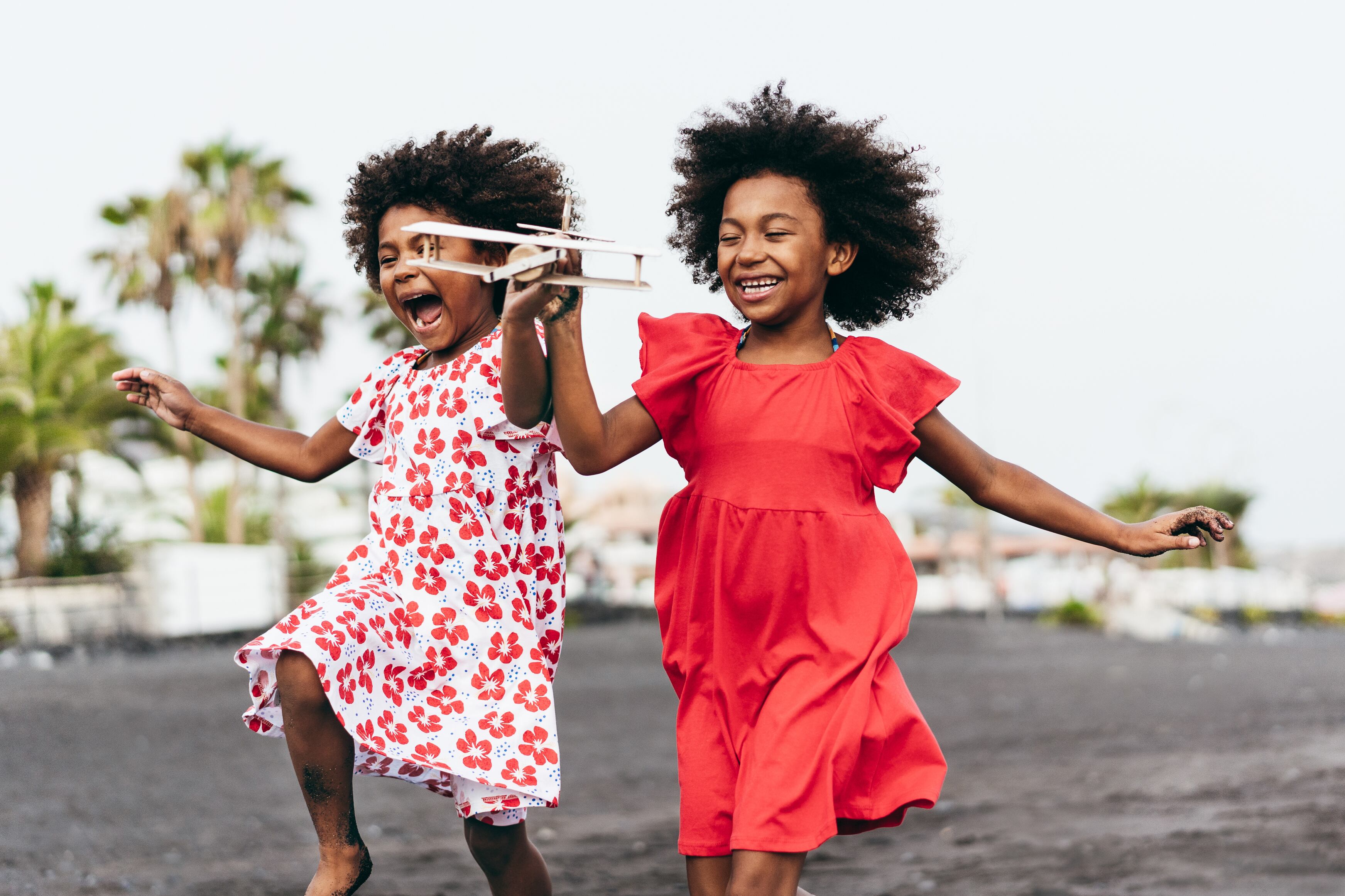 Hermanas gemelas afro corriendo en la playa mientras juegan con un avión de juguete de madera - Estilo de vida juvenil y concepto de viaje - Enfoque principal en la cara derecha del niño