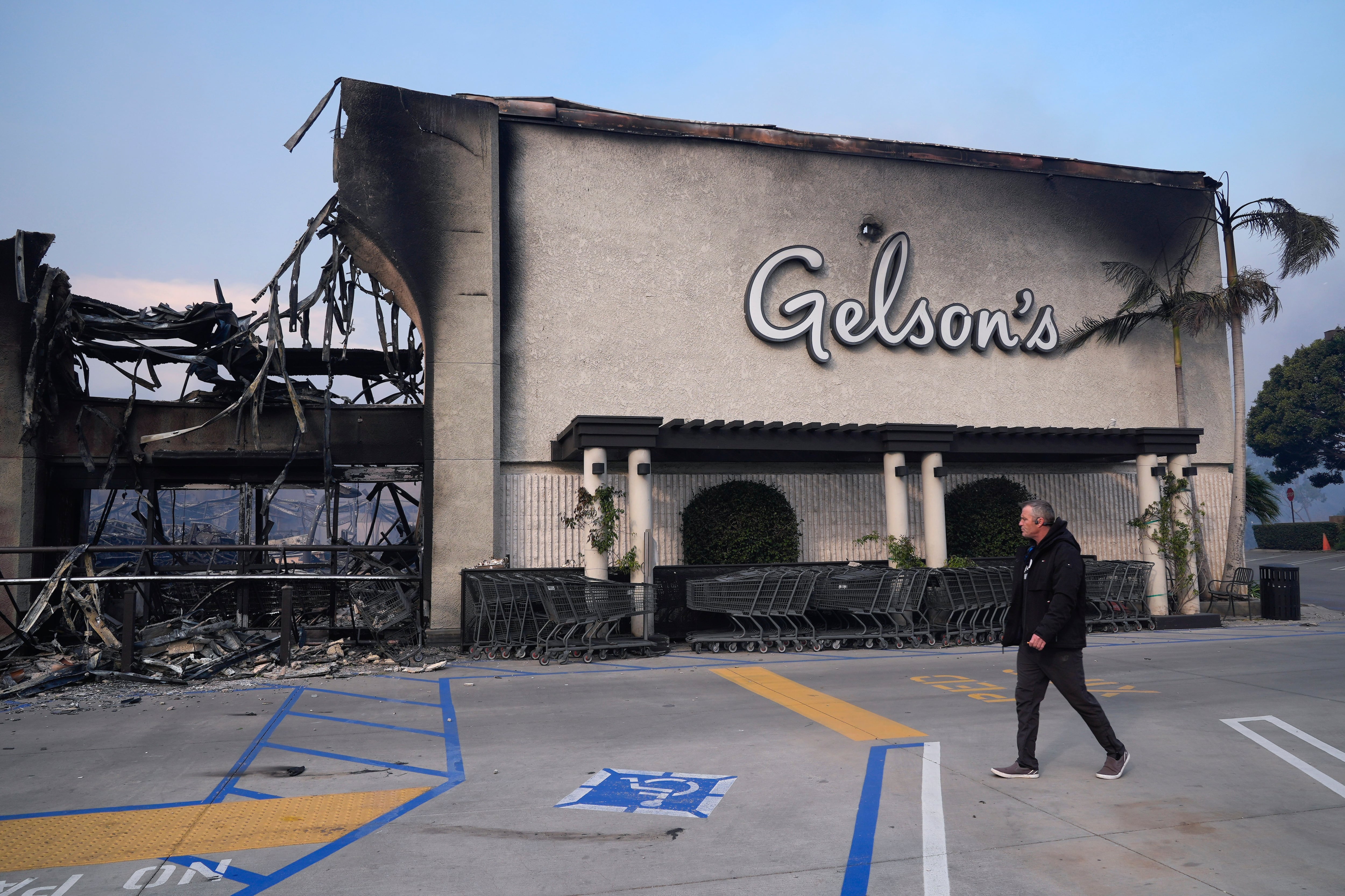 A man walks by Gelson's as the Palisades Fire ravages a neighborhood amid high winds in the Pacific Palisades neighborhood of Los Angeles, Wednesday, Jan. 8, 2025. (AP Photo/Damian Dovarganes)