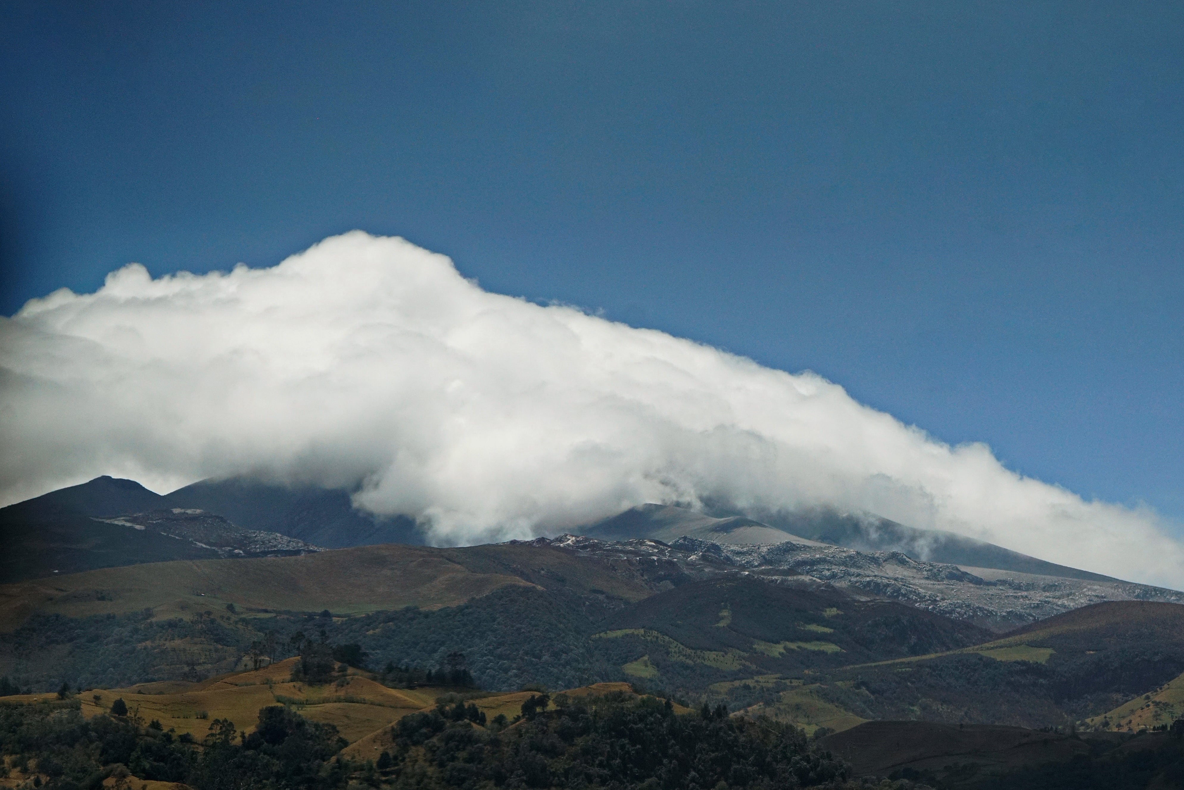 La constante actividad del volcán Puracé, ubicado en el departamento del Cauca, ha transformado drásticamente la vida de las comunidades indígenas y campesinas que habitan en sus alrededores. Este coloso, que forma parte del Parque Nacional Natural Puracé, ha mantenido en alerta máxima a la región debido a la constante emisión de gases, cenizas y material particulado, generando una crisis ambiental, económica y social que amenaza el sustento de miles de familias. Uno de los impactos más severos se registra en las fuentes hídricas. La contaminación de los ríos y quebradas de la zona con azufre y otros componentes químicos ha deteriorado la calidad del agua indispensable para el consumo humano y el riego. Esto no solo pone en riesgo la salud de los habitantes, sino que altera profundamente los ecosistemas locales, dejando a las comunidades en una situación de alta vulnerabilidad frente al desabastecimiento de agua limpia. La agricultura y la ganadería, principales motores económicos de la región, también sufren las consecuencias directas de este fenómeno natural. La caída persistente de ceniza ha provocado que baje drásticamente la producción de leche, ya que los pastizales se contaminan y el ganado padece problemas respiratorios y digestivos que merman su rendimiento. Paralelamente, la actividad volcánica afecta gravemente los cultivos de subsistencia y comerciales como la papa y las hortalizas, arruinando cosechas enteras bajo mantos de polvo gris. Ante este panorama, los pobladores del Puracé enfrentan el doble desafío de convivir con la constante amenaza de una erupción mayor y mitigar el desgaste diario de su economía. A pesar de los esfuerzos y monitoreos de las autoridades, el llamado de las comunidades locales es urgente: se requiere un apoyo integral que incluya asistencia técnica para el campo, sistemas alternativos de abastecimiento de agua y planes de contingencia económicos que les permitan resistir el embate de vivir a la sombra de un volcán activo.