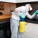 Professional exterminator in protective workwear spraying pesticide in apartment kitchen.