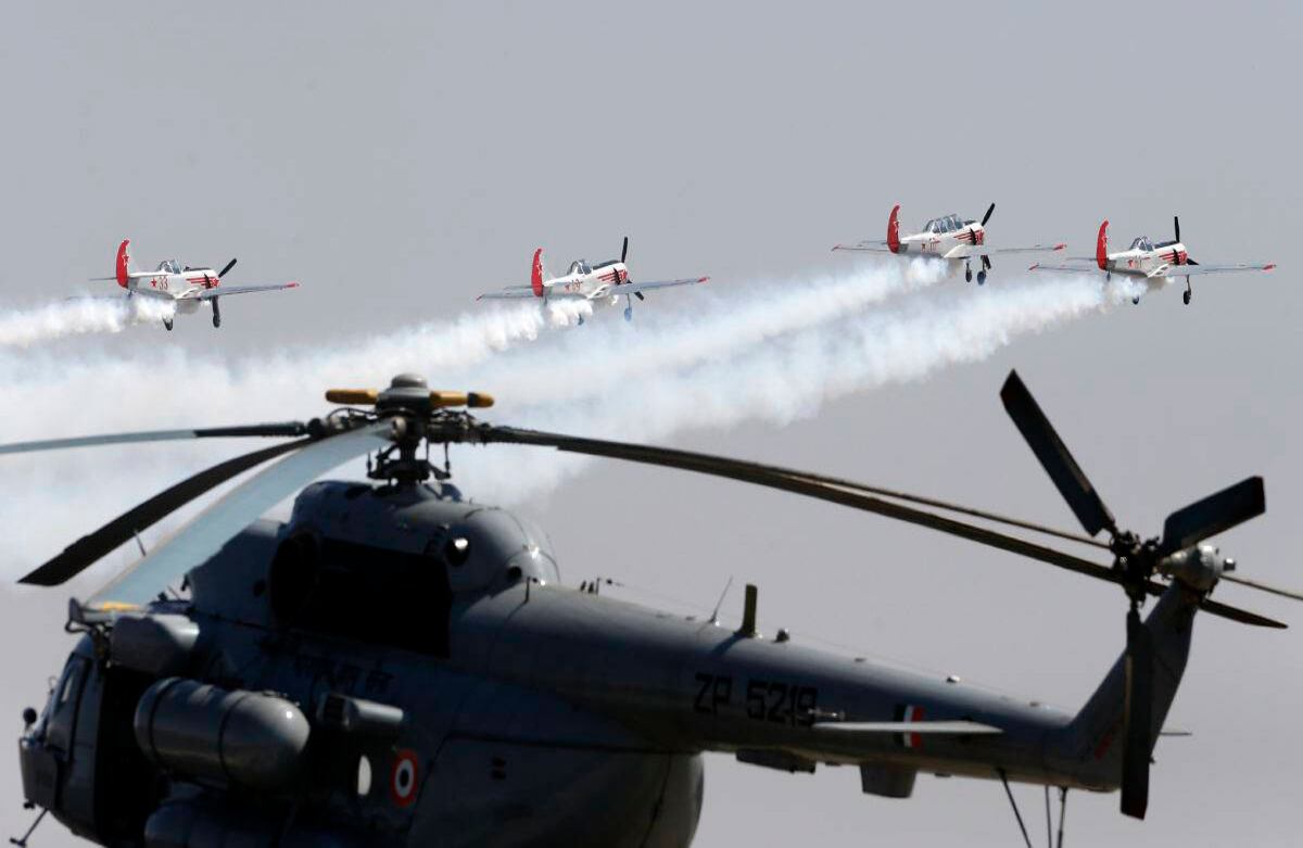 Los Yakovlevs, un equipo de acrobacia aérea con base en el Reino Unido que vuela aviones Yakovlev de diseño ruso, sobrevuelan un helicóptero de la Fuerza Aérea de la India en el tercer día de Aero India 2017 en la base aérea de Yelahanka en Bangalore, India, Jueves 16 de febrero de 2017. (AP Foto / Aijaz Rahi)  