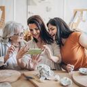 Multi-generation family women learning pottery art, they are modeling clay for making pottery in workshop, they are taking pictures of their work with smart phone.