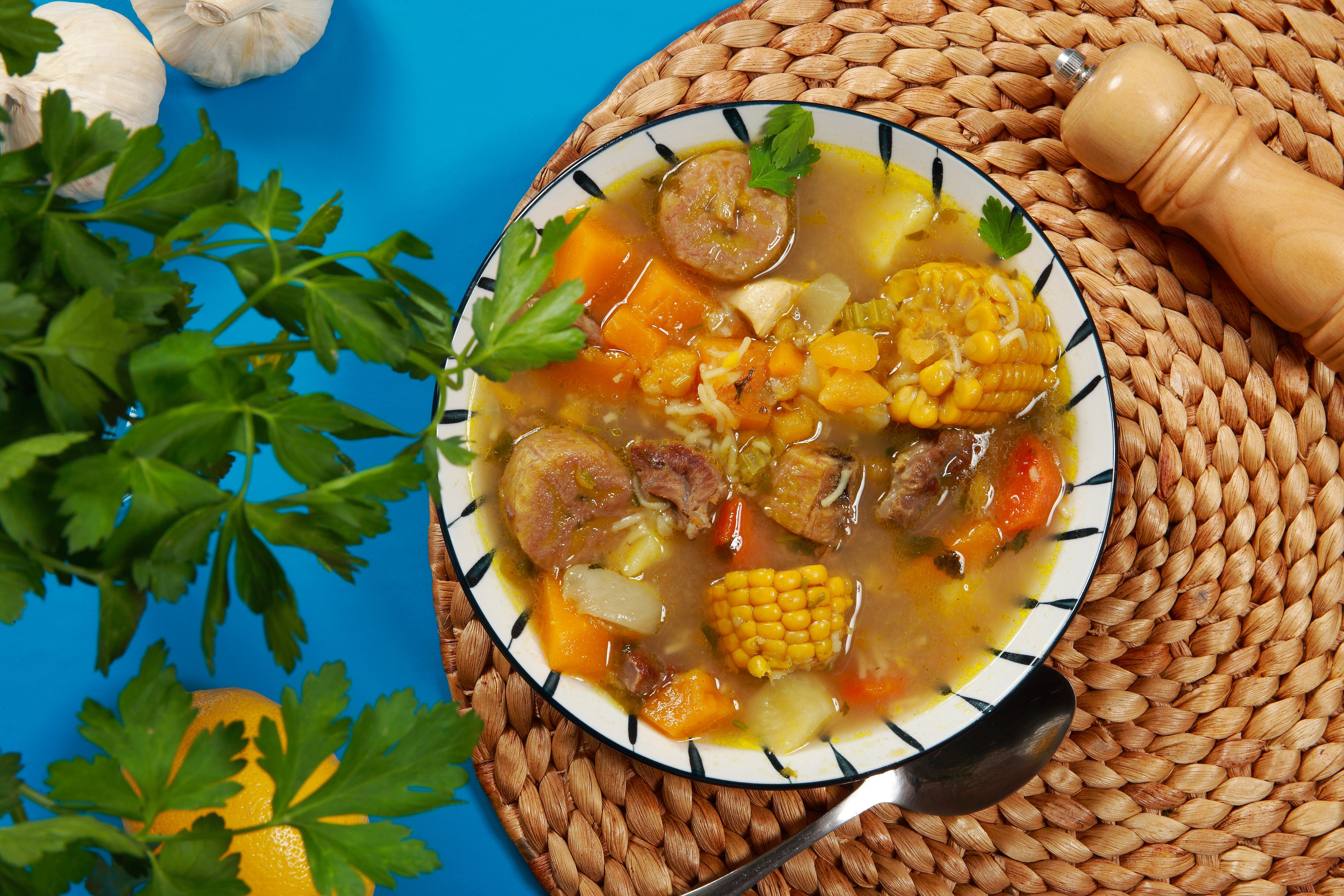 Sopa de estofado de carne tradicional colombiana que incluye batata de maíz, carne de res y pollo, cubierta con una salsa sabrosa y guarnición de arroz. Sancocho de Costilla sobre fondo azul.