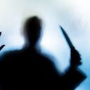 Backlit image of the silhouette of a person, probably a woman, wielding a sharp knife in an aggressive way. The silhouette is distorted, and the arms elongated, giving an alien-like quality. The image is sinister and foreboding, with an element of horror. The image conveys a domestic violence, knife crime theme. Horizontal image with copy space.