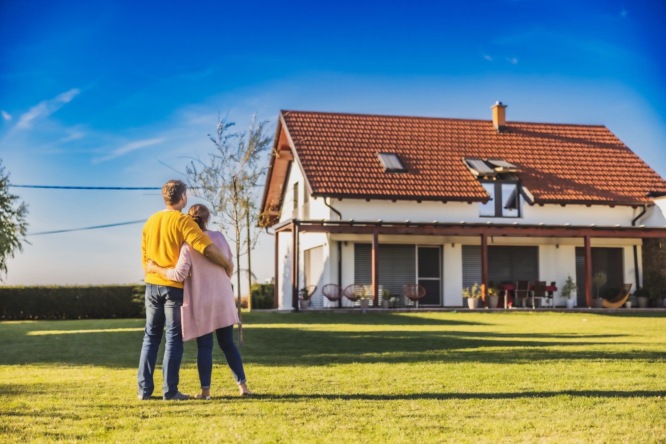 Couple,man with short brown hair and woman with long brown hair,hugging each other while standing on the lawn in the backyard of their new bought house,rear view,modern house in the background with almost clear sky in the evening