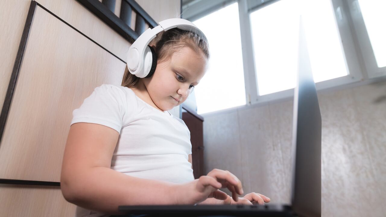 Una niña pequeña y gorda con una cara seria en los auriculares se sienta en el suelo en una habitación con una computadora portátil en los brazos y hace su tarea. Aprendizaje remoto