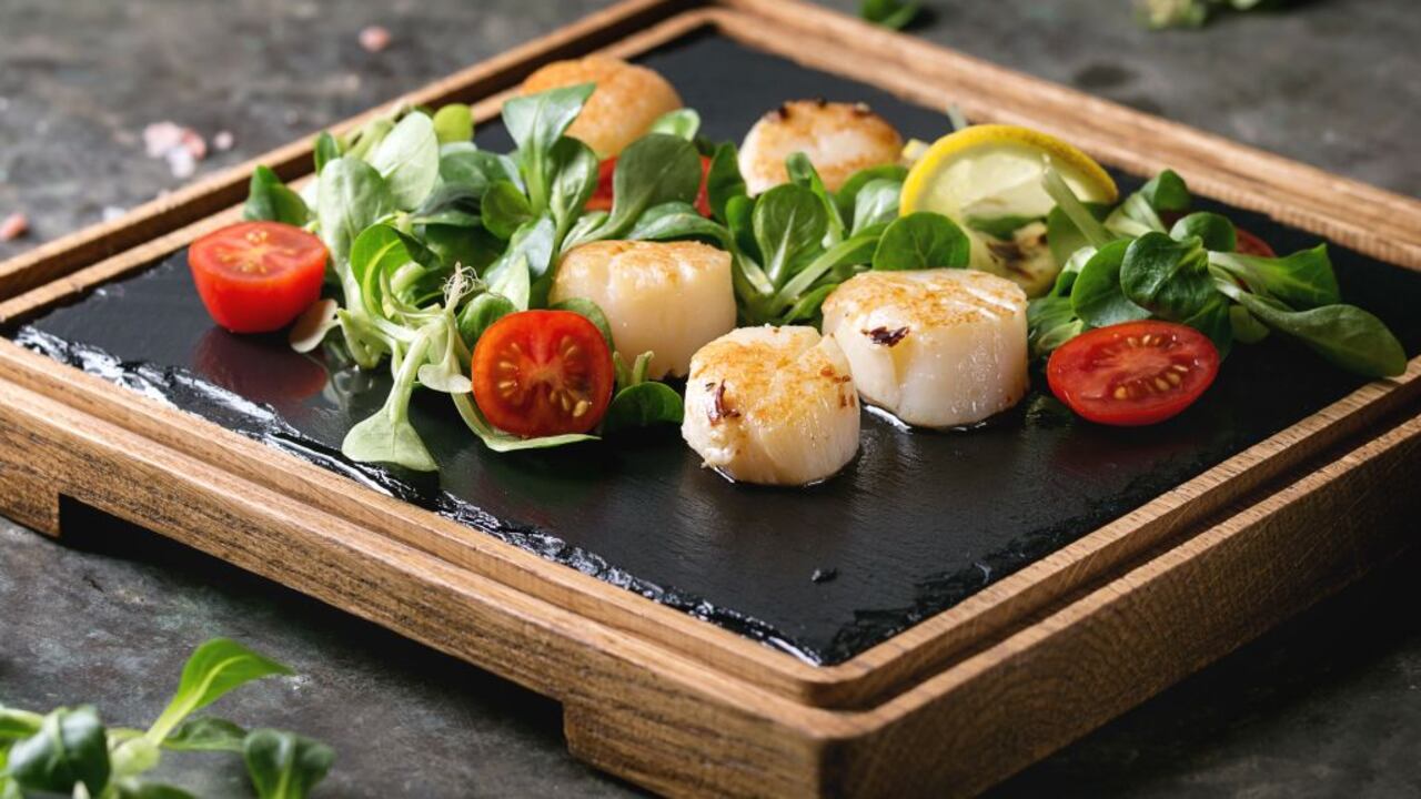 Vieiras fritas con limón, tomates cherry y ensalada verde servidas en una tabla de madera de pizarra negra sobre un viejo fondo de metal oscuro. De cerca.