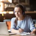 Mujer enfocada usando audífonos usando laptop en café, escribiendo notas, estudiante atractiva aprendiendo idioma, viendo seminarios web en línea, escuchando curso de audio, concepto de educación e-learning