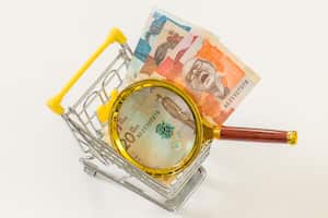 A miniature shopping cart with Colombian money and a magnifying glass. Economic and business concept, rising inflation and the country's economic upset. White background