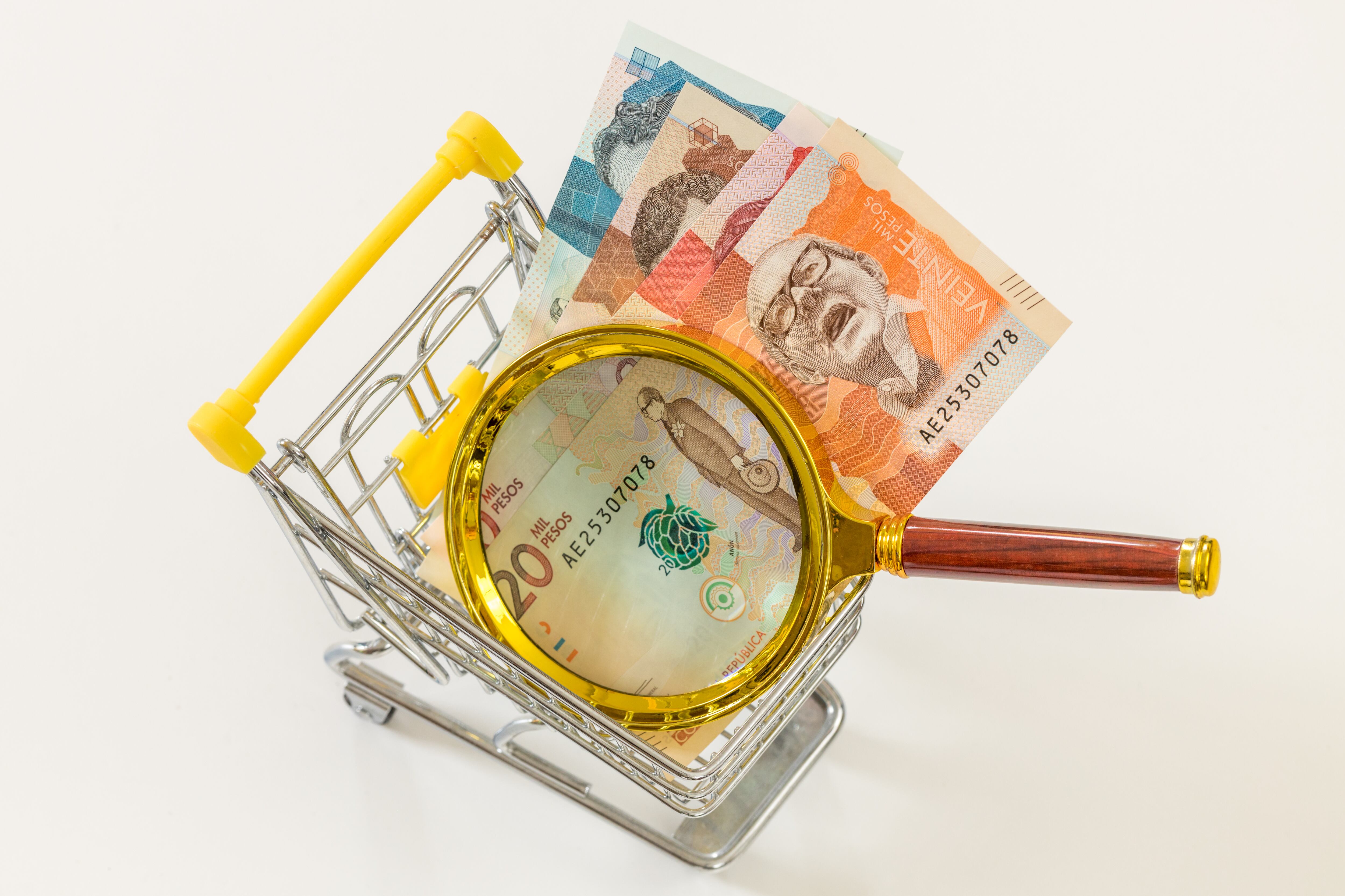 A miniature shopping cart with Colombian money and a magnifying glass. Economic and business concept, rising inflation and the country's economic upset. White background